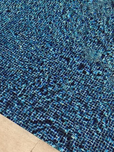 A close-up view of a swimming pool with a shimmering water surface. The pool is lined with small blue tiles that create a textured, mosaic pattern. The edge of the pool features a beige stone border, adding contrast to the vibrant blue water.