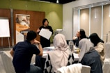 A group of people is engaged in a training session in a conference room setting. A presenter is standing and holding a sheet of paper, addressing the group seated at a round table. The attendees are actively listening, and there's a banner in the background advertising UX training. The room is well-lit, with a flipchart visible at one side.