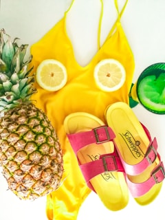 A yellow swimsuit is laid flat with two lemon slices placed as if they are part of the design. A fresh pineapple and a pair of yellow and pink sandals are arranged beside it. A green glass with a citrus slice is also present.
