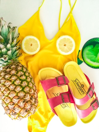 A yellow swimsuit is laid flat with two lemon slices placed as if they are part of the design. A fresh pineapple and a pair of yellow and pink sandals are arranged beside it. A green glass with a citrus slice is also present.