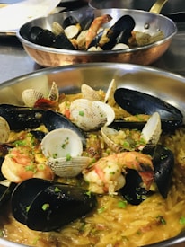 A close-up of a dish featuring a mix of seafood including mussels, clams, and shrimp atop a bed of seasoned orzo pasta. The seafood is garnished with fresh herbs, likely parsley, adding a touch of green to the golden brown tones of the pasta and shells. In the background, another similar dish is partially visible.
