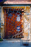 A wooden door adorned with several posters, framed by an artistic, blue, geometric metal gate. The gate features diagonal bars and intricate pipe-like structures. In front of the door, a wooden chair with metal legs is situated on the pavement.