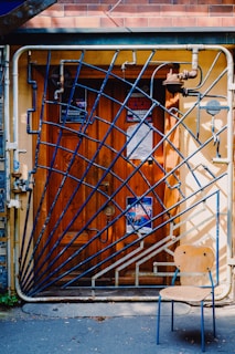 A wooden door adorned with several posters, framed by an artistic, blue, geometric metal gate. The gate features diagonal bars and intricate pipe-like structures. In front of the door, a wooden chair with metal legs is situated on the pavement.