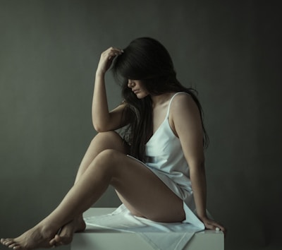 A woman with long dark hair is seated contemplatively on a white platform, wearing a light-colored slip dress. Her posture is introspective, with her head tilted downwards and one hand resting on her head. The background is a muted, dark tone, contributing to a somber, reflective ambiance.