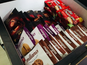 A box filled with various packaged snacks including chocolate chip cookies, branded as Milano, and confectioneries labeled as Lotte Choco Pie and Dark Fantasy. The packages are neatly arranged with an assortment of colors including red, purple, white, and black packaging.