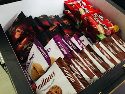 A box filled with various packaged snacks including chocolate chip cookies, branded as Milano, and confectioneries labeled as Lotte Choco Pie and Dark Fantasy. The packages are neatly arranged with an assortment of colors including red, purple, white, and black packaging.