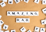 Letter tiles spell out the words 'AMAZING DAD' on a tan surface, surrounded by additional scattered tiles featuring random letters.