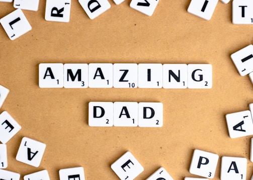 Letter tiles spell out the words 'AMAZING DAD' on a tan surface, surrounded by additional scattered tiles featuring random letters.