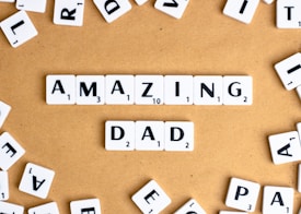 Letter tiles spell out the words 'AMAZING DAD' on a tan surface, surrounded by additional scattered tiles featuring random letters.