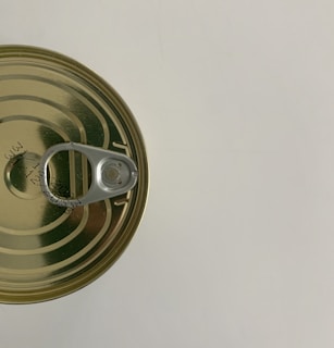 A top view of a sealed, metallic tin can with a pull-tab opener. The can is placed on a light surface and has some printed text or numbers on its top surface.
