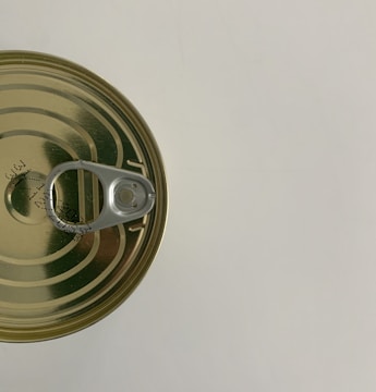 A top view of a sealed, metallic tin can with a pull-tab opener. The can is placed on a light surface and has some printed text or numbers on its top surface.