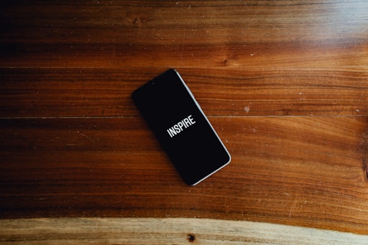 A smartphone with a black screen displaying the word 'INSPIRE' in white text is placed on a smooth wooden surface with visible grain patterns.