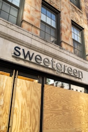 A boarded-up storefront of a building with the sign 'sweetgreen' displayed above. The structure has a classic architectural style with large windows and decorative elements. The wooden boards cover the lower part of the entrance, suggesting potential renovation or protection.