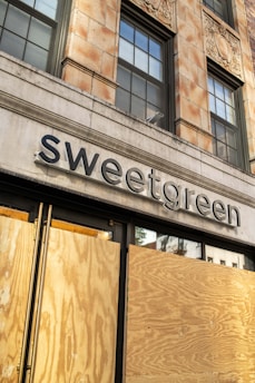 A boarded-up storefront of a building with the sign 'sweetgreen' displayed above. The structure has a classic architectural style with large windows and decorative elements. The wooden boards cover the lower part of the entrance, suggesting potential renovation or protection.