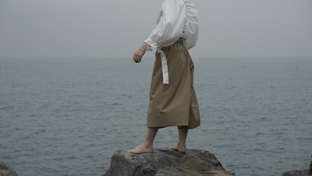woman in white hijab and brown dress standing on rock near body of water during daytime