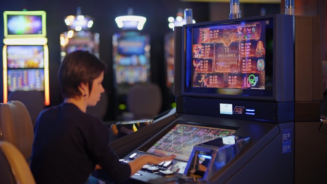 A person is seated at a slot machine console in a casino, focusing intently on the game displayed on the screen. The background shows several other illuminated gaming machines, adding a vibrant and colorful atmosphere to the scene.