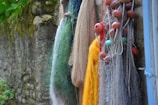 A collection of fishing nets of various colors hang against a rustic stone wall. The nets are adorned with red and turquoise floats, featuring shades of yellow, green, blue, and brown threads. Some parts of the wall have moss and plants, adding a natural touch.