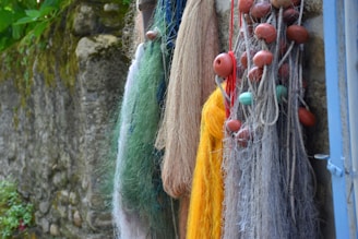 A collection of fishing nets of various colors hang against a rustic stone wall. The nets are adorned with red and turquoise floats, featuring shades of yellow, green, blue, and brown threads. Some parts of the wall have moss and plants, adding a natural touch.