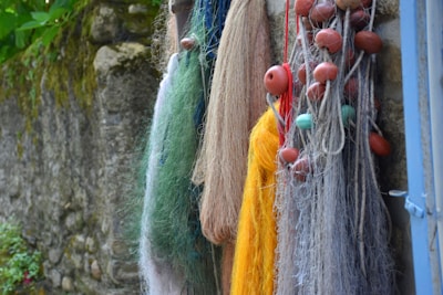 A collection of fishing nets of various colors hang against a rustic stone wall. The nets are adorned with red and turquoise floats, featuring shades of yellow, green, blue, and brown threads. Some parts of the wall have moss and plants, adding a natural touch.