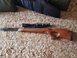 A wooden rifle with a scope is placed on a textured carpet. The rifle has a sleek design and the stock is made of polished wood.