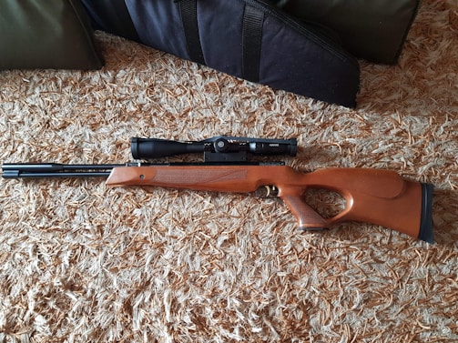 A wooden rifle with a scope is placed on a textured carpet. The rifle has a sleek design and the stock is made of polished wood.