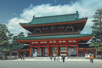 A traditional Japanese temple with a green, intricately tiled roof and white and red walls. The large entrance has multiple stories and is surrounded by tall pine trees. Several people in both modern and traditional clothing are walking and standing around the temple.