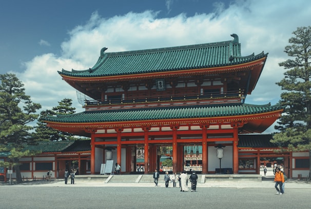 A traditional Japanese temple with a green, intricately tiled roof and white and red walls. The large entrance has multiple stories and is surrounded by tall pine trees. Several people in both modern and traditional clothing are walking and standing around the temple.