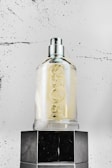 A clear glass bottle with a spray nozzle is sitting on top of a glossy black surface. The bottle is marked with embossed lettering. The background is a textured, speckled white surface, adding a modern and minimalist touch to the scene.