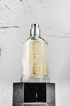 A clear glass bottle with a spray nozzle is sitting on top of a glossy black surface. The bottle is marked with embossed lettering. The background is a textured, speckled white surface, adding a modern and minimalist touch to the scene.
