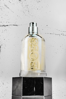 A clear glass bottle with a spray nozzle is sitting on top of a glossy black surface. The bottle is marked with embossed lettering. The background is a textured, speckled white surface, adding a modern and minimalist touch to the scene.