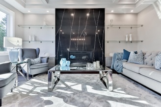 A contemporary living room featuring a black marble accent wall with subtle veining and a modern fireplace at its center. The room is furnished with a large gray sofa adorned with patterned and solid blue cushions, and a pair of gray upholstered armchairs. A glass-topped coffee table with geometric metallic legs holds decorative items including blue vases and candles. Glass shelves, positioned on either side of the fireplace, display minimalistic decor. Natural light streams in from a window on the left, enhancing the room’s bright and airy ambiance.