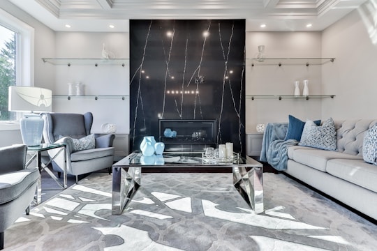 A contemporary living room featuring a black marble accent wall with subtle veining and a modern fireplace at its center. The room is furnished with a large gray sofa adorned with patterned and solid blue cushions, and a pair of gray upholstered armchairs. A glass-topped coffee table with geometric metallic legs holds decorative items including blue vases and candles. Glass shelves, positioned on either side of the fireplace, display minimalistic decor. Natural light streams in from a window on the left, enhancing the room’s bright and airy ambiance.