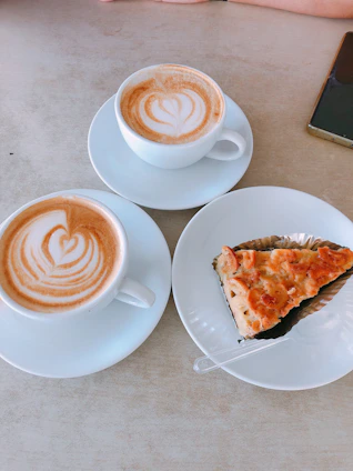 Two cups of coffee with latte art designs and a slice of pizza on a white plate. There is a smartphone on the table and a person's arm visible at the top of the image.