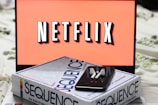 A screen displaying the Netflix logo in bold, black and white lettering against a vibrant red background. In front of the screen, there is a closed box for the board game 'Sequence', with a smartphone and wireless earbuds resting on top.