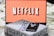 A screen displaying the Netflix logo in bold, black and white lettering against a vibrant red background. In front of the screen, there is a closed box for the board game 'Sequence', with a smartphone and wireless earbuds resting on top.