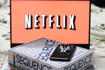 A screen displaying the Netflix logo in bold, black and white lettering against a vibrant red background. In front of the screen, there is a closed box for the board game 'Sequence', with a smartphone and wireless earbuds resting on top.
