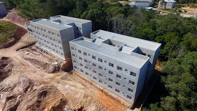 Two large, rectangular concrete buildings with multiple windows are situated in a partially cleared area surrounded by dense green forest. The buildings are under construction, with visible construction materials and unfinished ground work.
