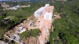 Aerial view of a construction site surrounded by dense green forest. The site includes several multi-story buildings in various stages of completion and construction vehicles. Temporary structures and materials are scattered across the area, with a dirt road running through the site.