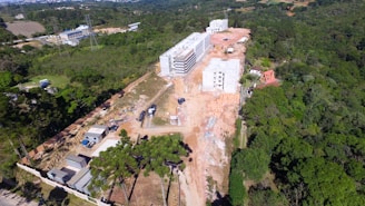 Aerial view of a construction site surrounded by dense green forest. The site includes several multi-story buildings in various stages of completion and construction vehicles. Temporary structures and materials are scattered across the area, with a dirt road running through the site.