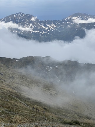 Hikers traverse a mist-covered mountain slope with snow-capped peaks looming in the background. The landscape is rugged and expansive, with clouds adding a dramatic atmosphere to the scene.