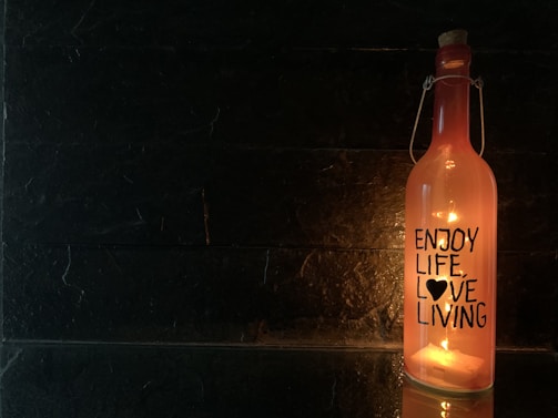 A glass bottle with a cork stopper, illuminated from within by a warm light. The bottle is inscribed with the words 'ENJOY LIFE, LOVE LIVING' with a heart symbol. The background is a dark, textured surface, creating a stark contrast with the glowing bottle.