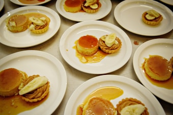 Multiple white plates are arranged on a table, each holding desserts comprised of caramel flan and banana-topped tartlets. The flan is glistening with a caramel sauce, and the tartlets feature banana slices on top, with a swirl of cream filling visible.