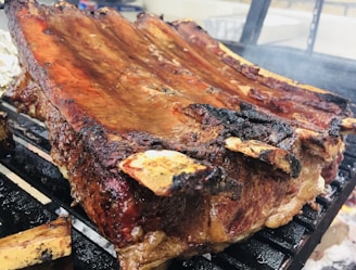 A large rack of ribs is being grilled, showcasing a beautifully charred exterior with a glossy sheen of sauce. The ribs are placed over a hot grill, with some smoke visible, suggesting they're being cooked over an open flame.