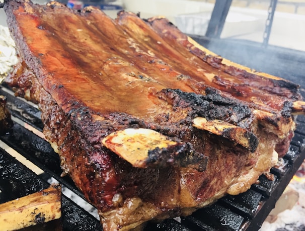 A large rack of ribs is being grilled, showcasing a beautifully charred exterior with a glossy sheen of sauce. The ribs are placed over a hot grill, with some smoke visible, suggesting they're being cooked over an open flame.