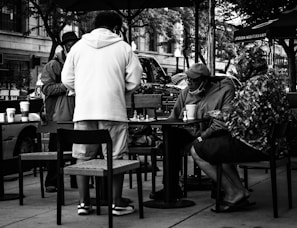 A black and white scene captures several people gathered around a small outdoor table, engaged in playing a game of chess. The individuals are casually dressed and appear to be enjoying a relaxed moment. Coffee cups are scattered on the table and chairs, suggesting a laid-back cafe environment. The background features parked cars and trees, indicating an urban setting.