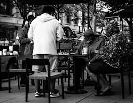 A black and white scene captures several people gathered around a small outdoor table, engaged in playing a game of chess. The individuals are casually dressed and appear to be enjoying a relaxed moment. Coffee cups are scattered on the table and chairs, suggesting a laid-back cafe environment. The background features parked cars and trees, indicating an urban setting.