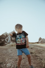 A young child with shoulder-length hair stands on a rocky path in a natural landscape. The child is wearing a black t-shirt with cartoon characters and 'Disney Crew' written on it, along with blue shorts and checkered shoes. The surroundings include dry grass and hills under a clear blue sky.