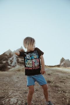 A young child with shoulder-length hair stands on a rocky path in a natural landscape. The child is wearing a black t-shirt with cartoon characters and 'Disney Crew' written on it, along with blue shorts and checkered shoes. The surroundings include dry grass and hills under a clear blue sky.
