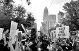 A large crowd of people is gathered holding signs for a Black Lives Matter protest in an urban setting. The image shows diverse participants, some are wearing masks, and one individual is carrying a child. Tall buildings and trees are visible in the background.