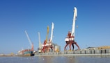 Large port cranes stand tall by the waterfront, with a cargo ship docked nearby. The sky is a clear blue, setting a contrasting backdrop to the industrial scene. The surrounding area includes warehouses and storage facilities, while seagulls hover above the water.